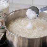 A tablespoon of organic coconut oil being dropped into boiling water with white rice in a pot